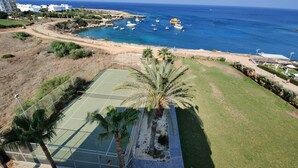 A tennis court with a palm tree, a beach, and boats in the background. - Charming 1-bed Apartment in Protaras, Cyprus (Paralimni)
