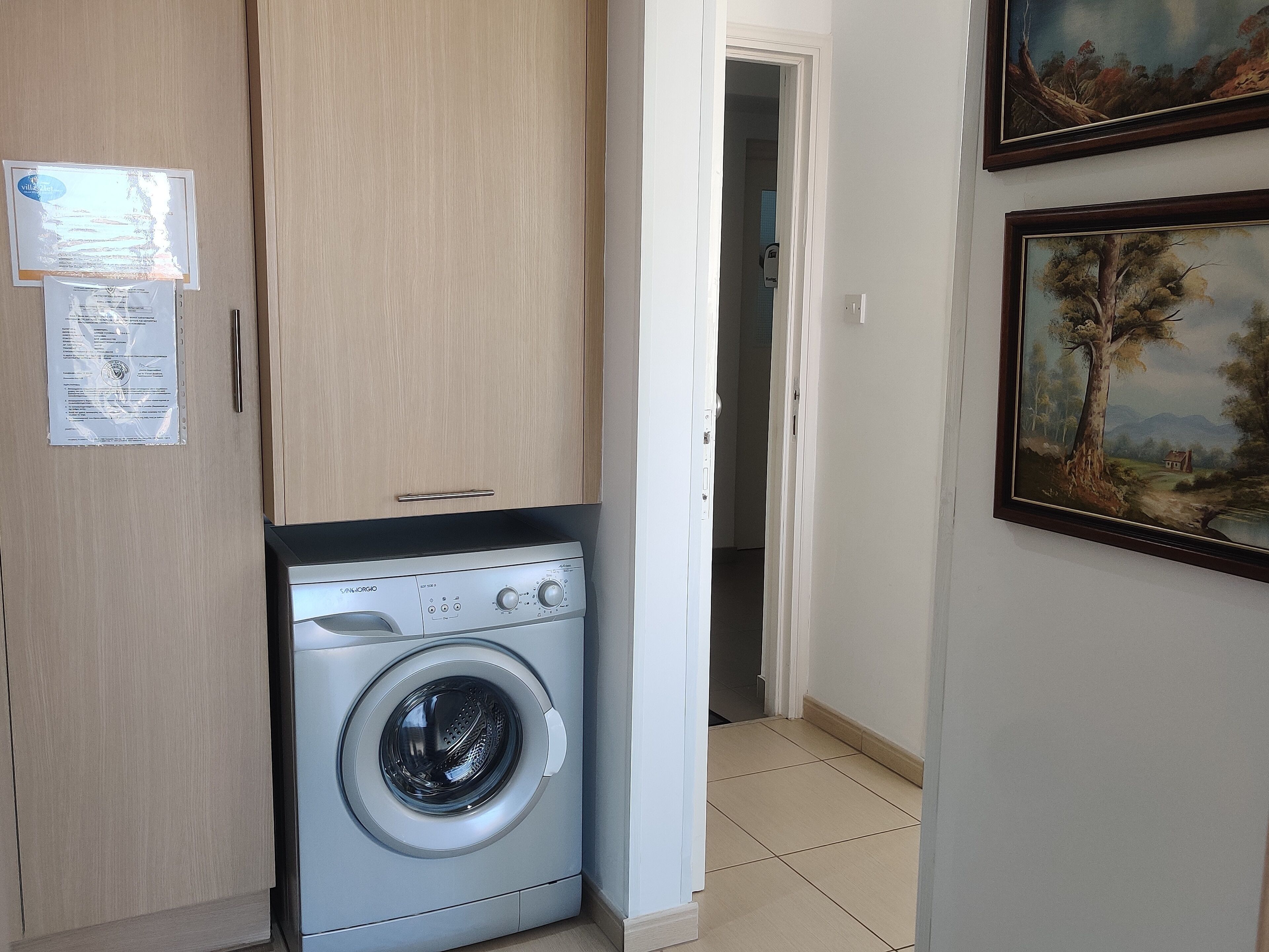 A laundry room with a front-loading washing machine, wooden cabinets, and a door leading to another room.