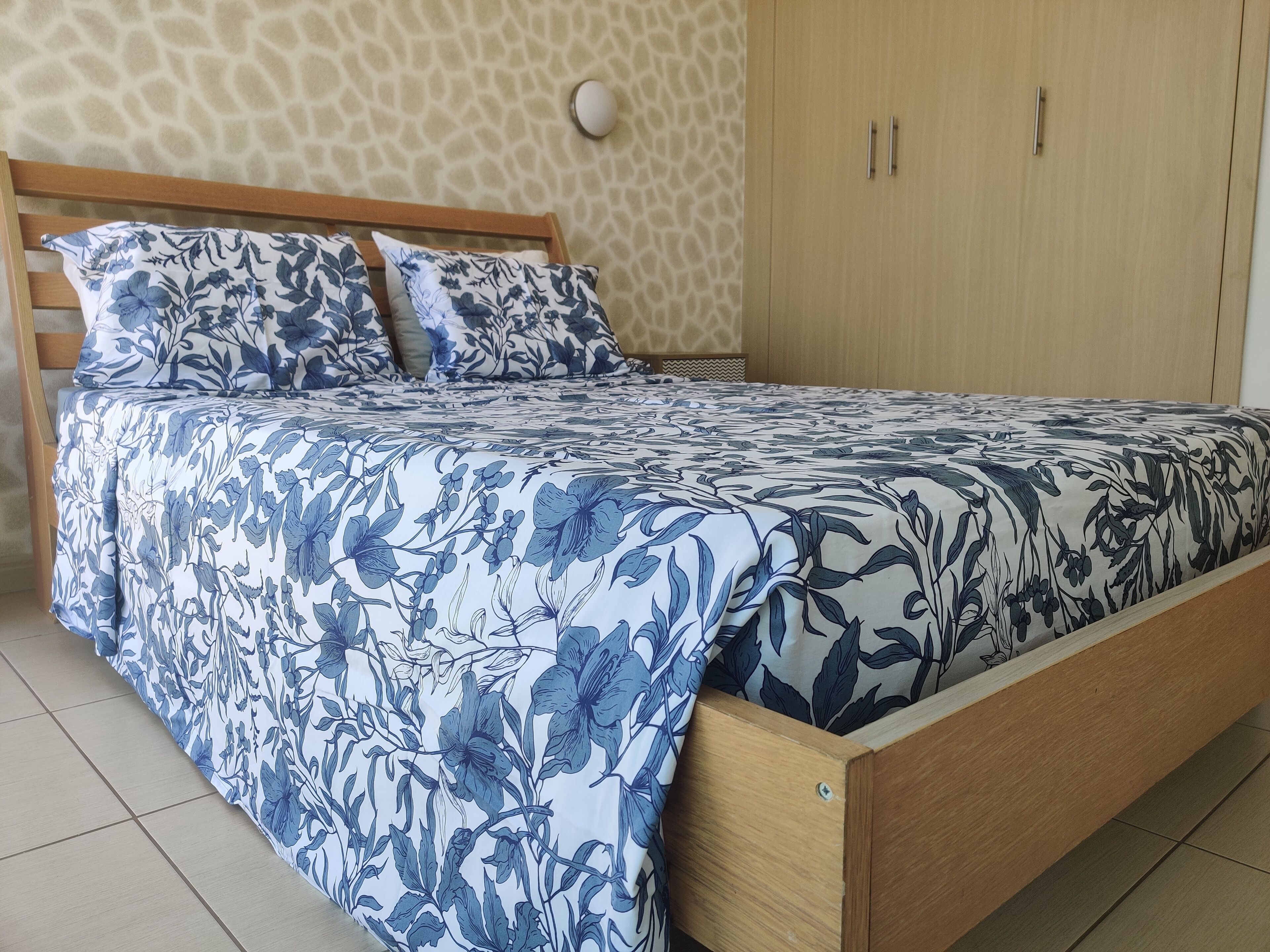 Una cama con colcha y almohadas estampadas con motivos florales, cabecera de madera y pared de color claro con textura.