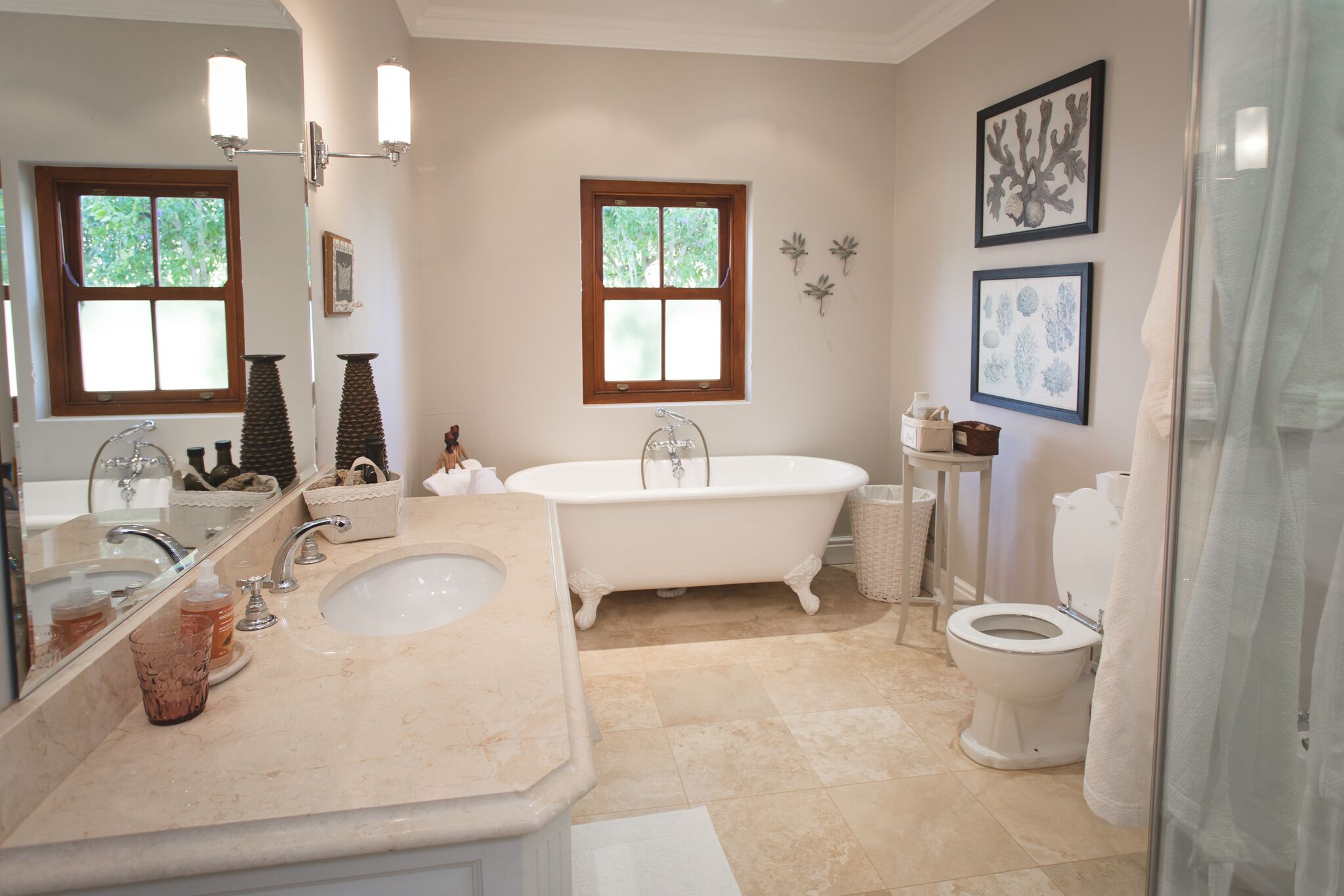 A bathroom with a freestanding bathtub, a sink, and a toilet. There are two windows providing natural light, and the walls are decorated with framed artwork.