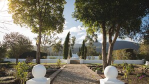 A garden pathway leading to a white structure with spherical sculptures at the ends, surrounded by trees and a view of distant hills.