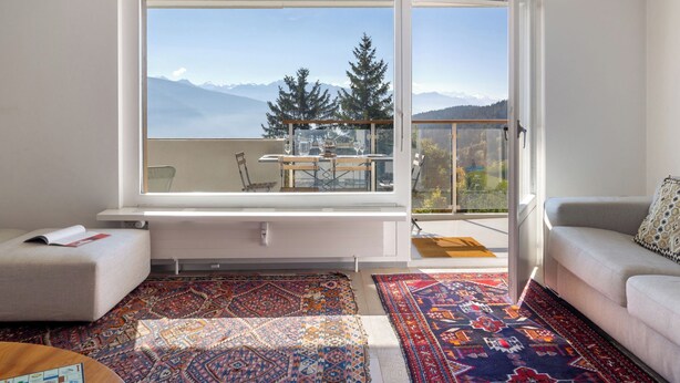 Uma sala de estar moderna com uma janela grande que oferece vista para as montanhas, um sofá, uma mesa de centro e um tapete estampado.