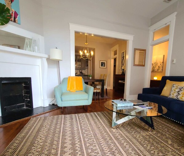 Una sala de estar con chimenea, un sillón azul, una mesa de centro de vidrio y un sofá azul con alfombra estampada.