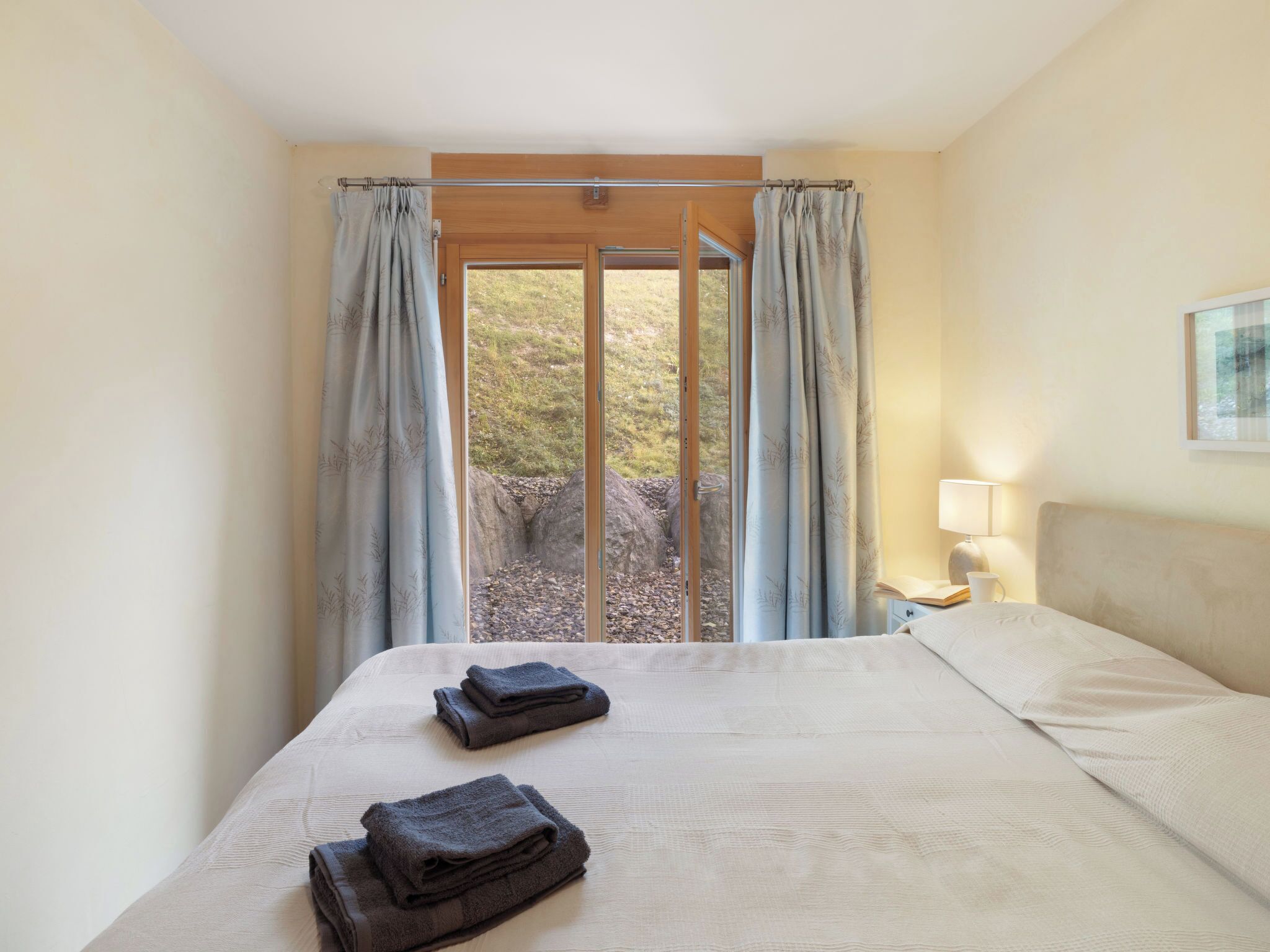 Hotelkamer met een bed, nachtkastje, lamp en uitzicht op een rotsachtig landschap door een glazen deur.