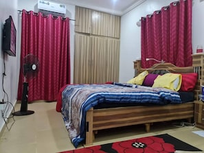A bedroom with a wooden bed, red curtains, a standing fan, and a tiled floor. - Smart 3 bed Apartment With Fast Wi-fi & 24hr Light (Abuja)