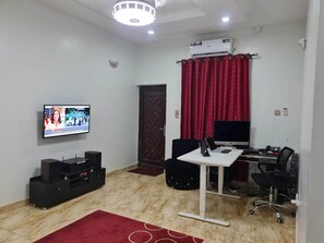 A room with a TV, a desk with a computer, a chair, a red curtain, and an air conditioner. - Smart 3 bed Apartment With Fast Wi-fi & 24hr Light (Abuja)