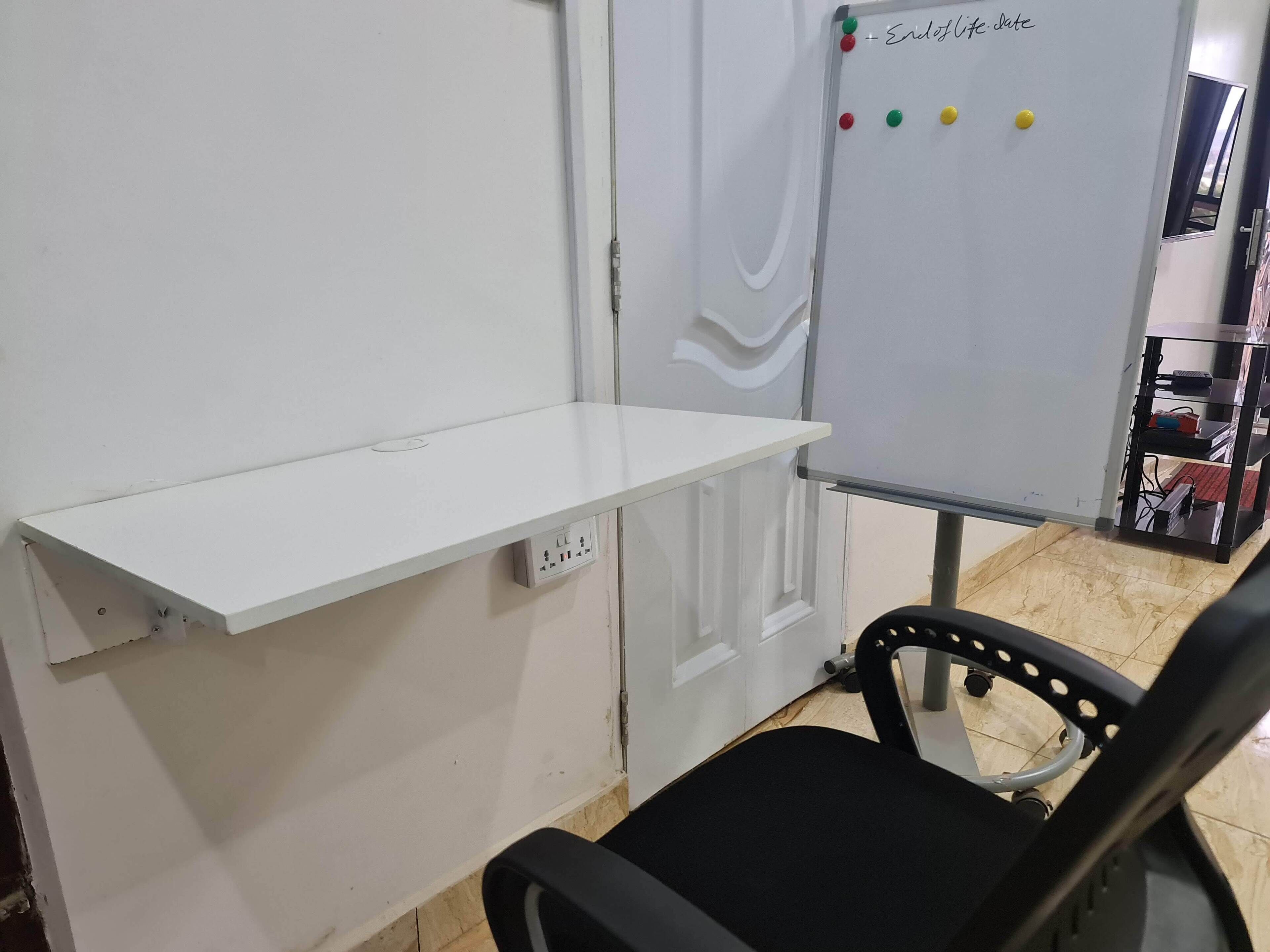 A room with a white desk, a black chair, a whiteboard with colored markers, and a shelf with items.