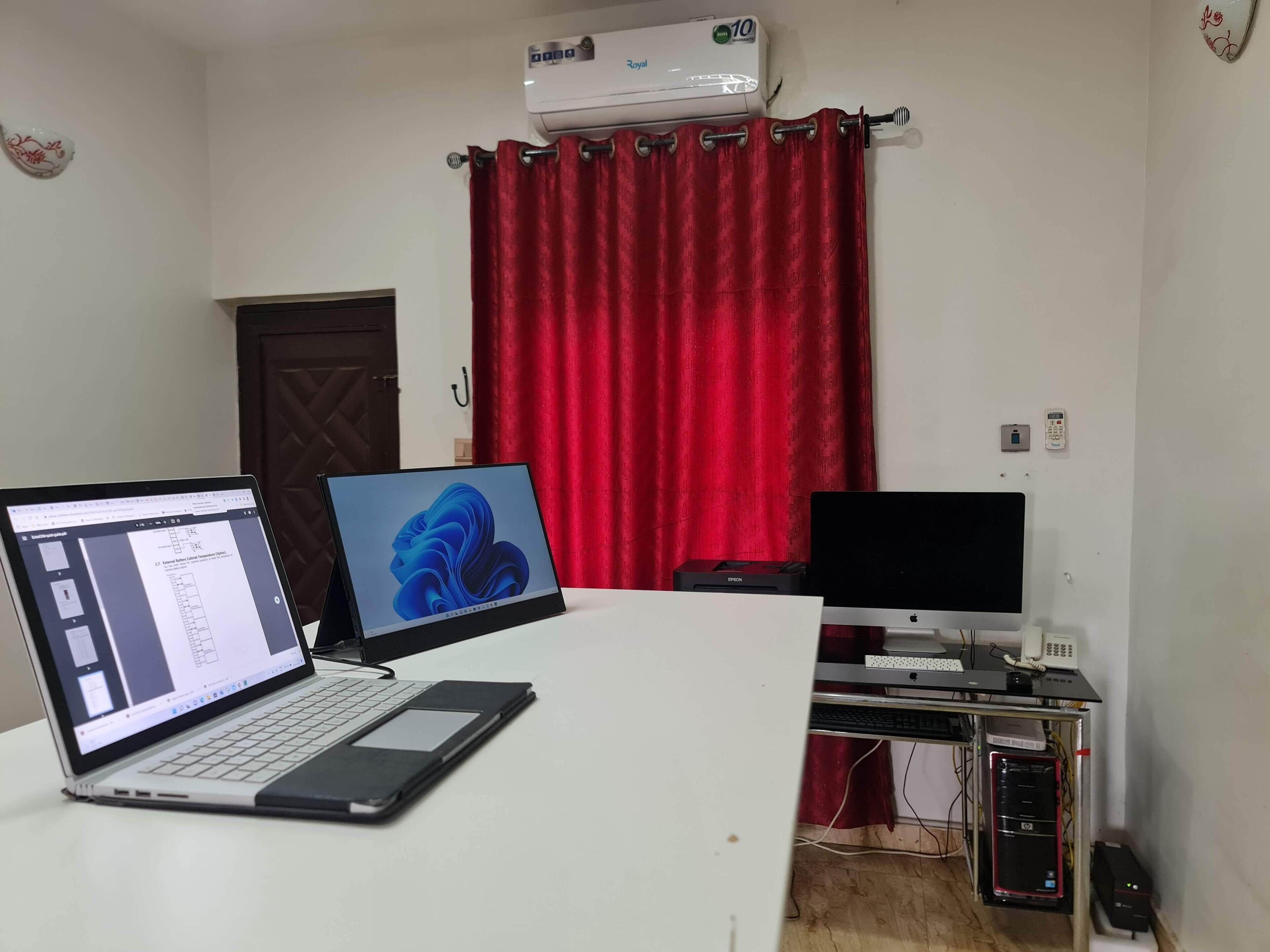A room with a desk, two laptops, a desktop monitor, an air conditioner, and a red curtain.