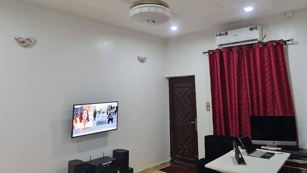 A room with a ceiling fan, an air conditioning unit, a television mounted on the wall, a desk with a laptop, a chair, and a door with a decorative pattern. - Smart 3 bed Apartment With Fast Wi-fi & 24hr Light (Abuja)