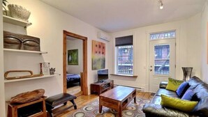 A living room with a black leather sofa, wooden coffee table, and a patterned rug. There are shelves with decorative items and a television on a stand. - Luxury Quiet Old Montreal Home (Montréal)