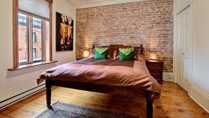 A bedroom with a brick wall, a bed with a wooden headboard, a nightstand, and a window with blinds. - Luxury Quiet Old Montreal Home (Montréal)