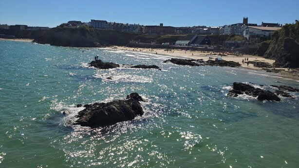 Paesaggio costiero con acque rocciose, una spiaggia sabbiosa e edifici sullo sfondo.