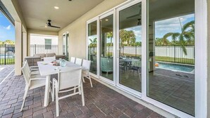Un patio techado con mesa y sillas, ventilador de techo y vista a una piscina y palmeras.