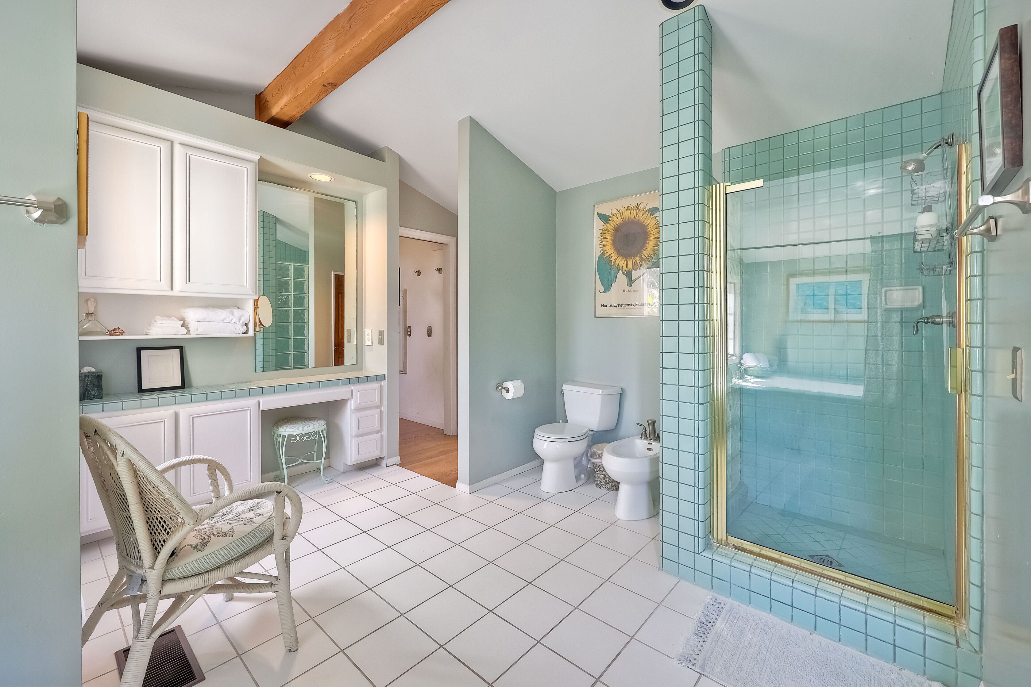 Ein Badezimmer mit weißem Waschbecken, zwei Toiletten, einer Duschkabine mit Glastür und einem Korbsessel.