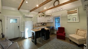 A compact kitchen with white cabinets, a refrigerator, and a microwave. There is a dining area with a table and chairs, and a red armchair. The ceiling has wooden beams and a ceiling fan.