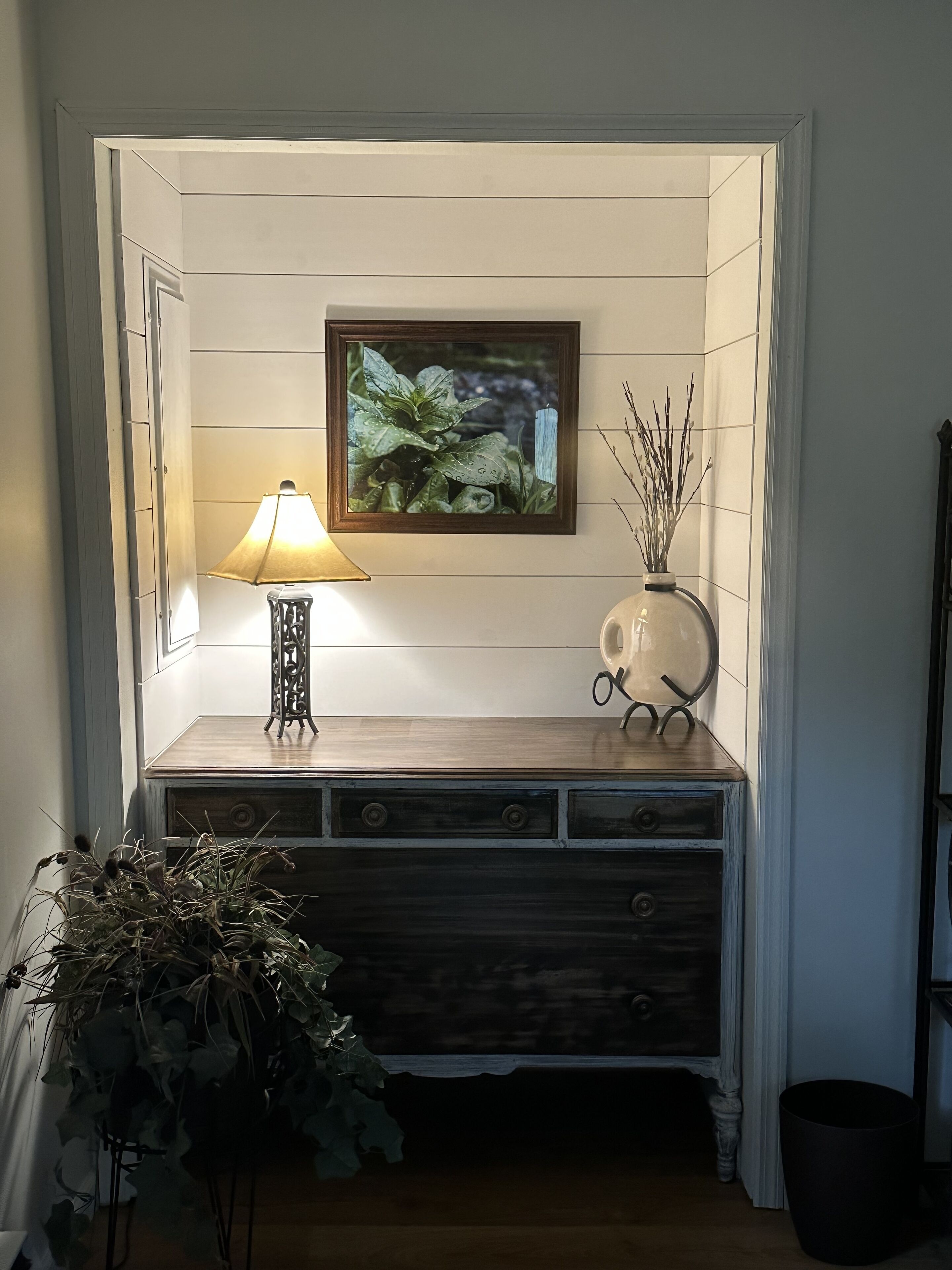 Ett litet, avskilt sittområde med en träbänk, ett bord med en lampa, en inramad tavla och en vas med grenar.