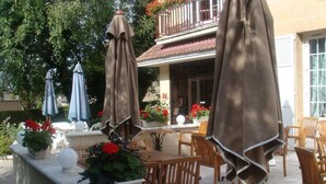 Een terras buiten met houten stoelen en tafels, parasols, potplanten en een balkon met rode bloemen.