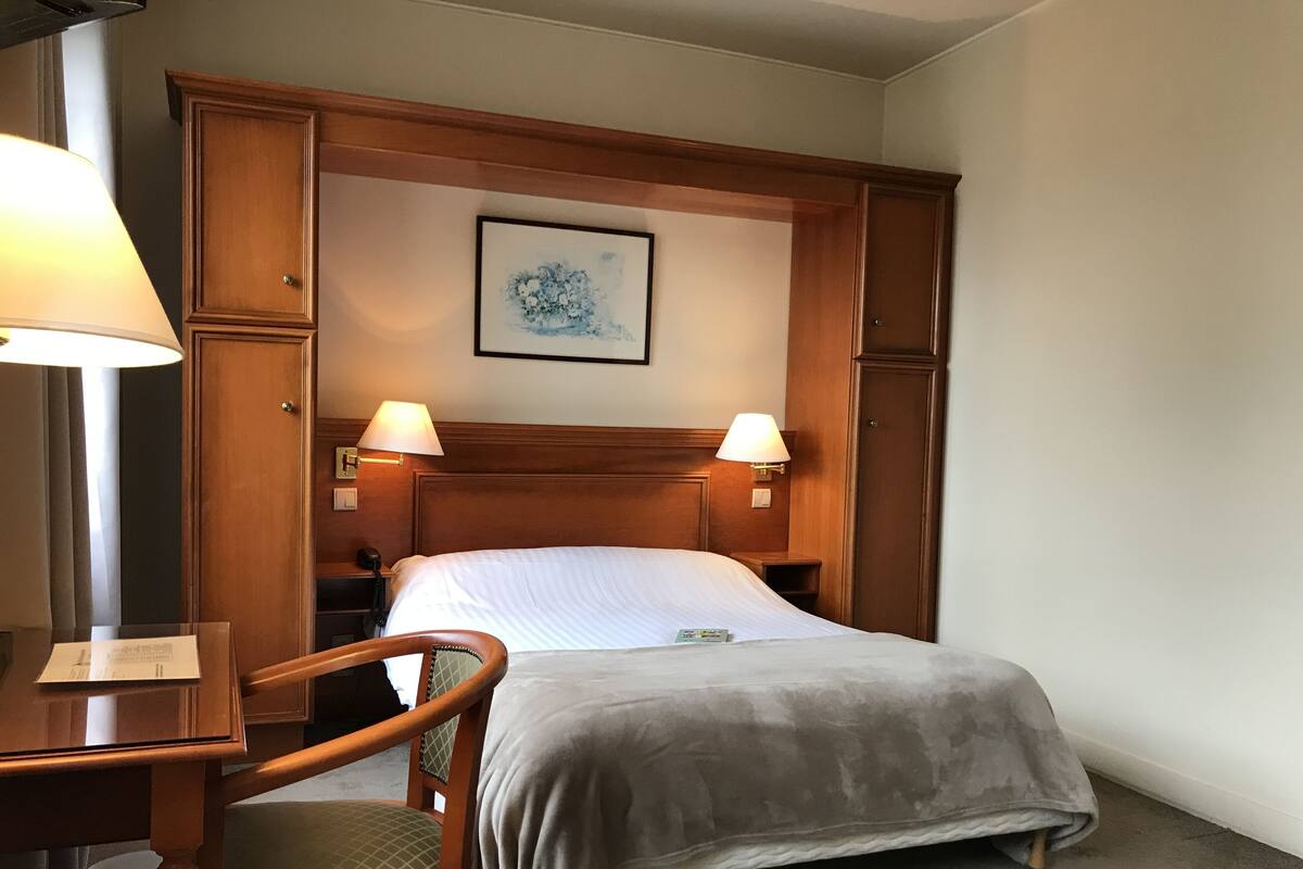 A hotel room with a wooden headboard, two bedside lamps, a bed with a grey blanket, a wooden chair, and a framed picture on the wall.