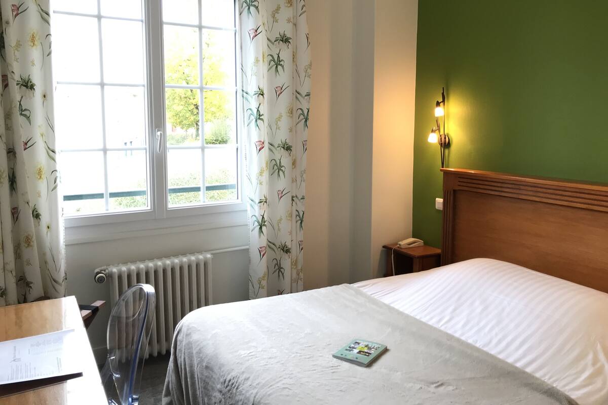 A hotel room with a bed, a desk, a chair, a radiator, and a window with curtains.