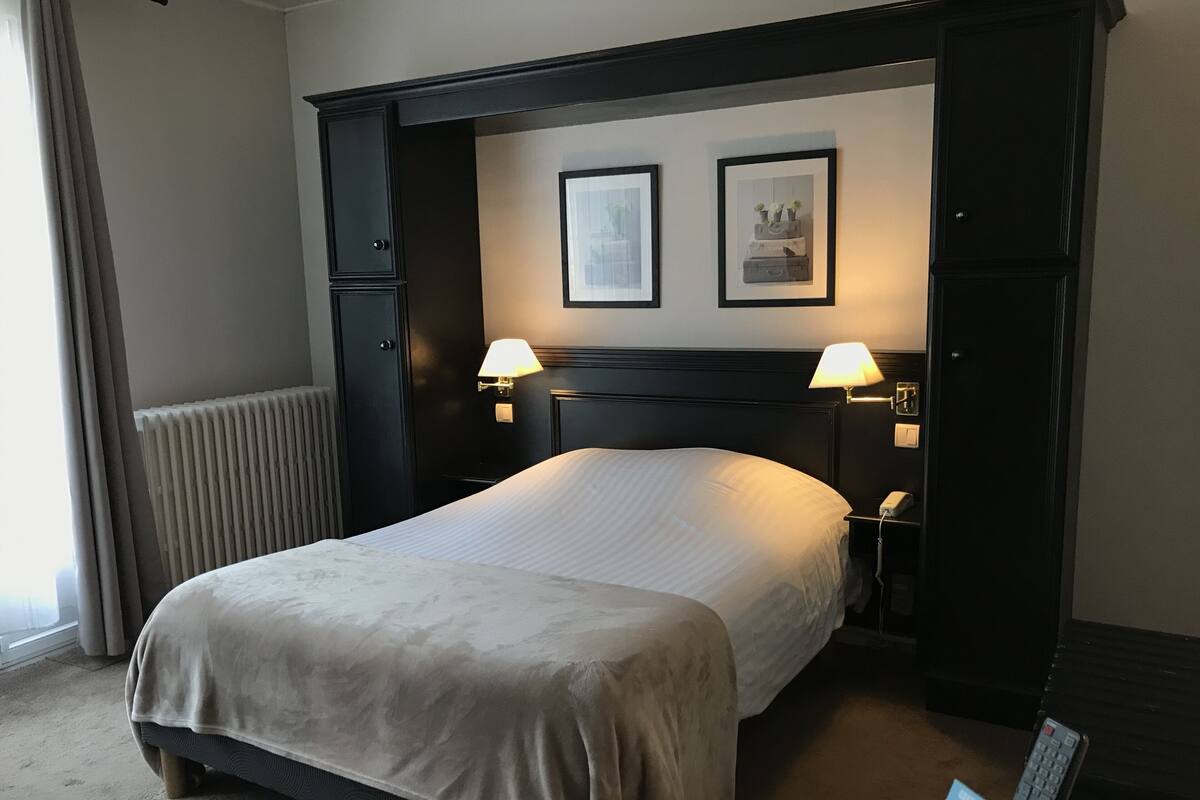 A hotel room with a bed, two bedside lamps, a nightstand, a telephone, a radiator, and two framed pictures on the wall.