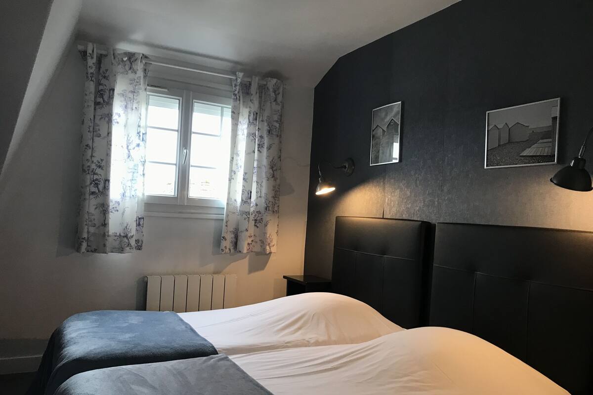 A bedroom with two beds, a window with curtains, a radiator, and two wall-mounted lamps.