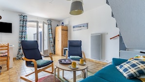 A modern living room with a blue sofa, wooden chairs, a small round table with cups and a tray, a wooden cabinet, and a television on the wall.