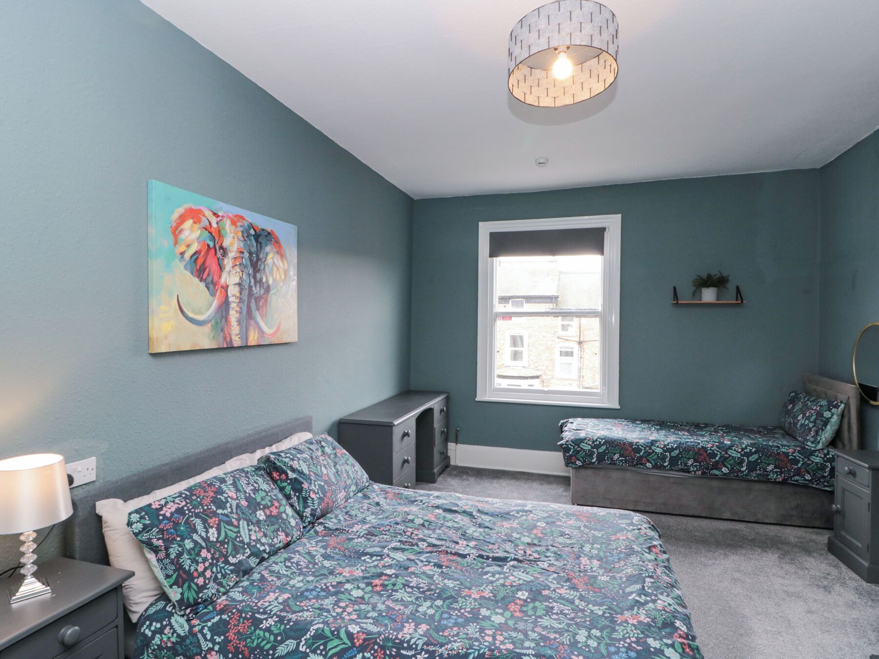 A bedroom with teal walls, a bed with floral bedding, a nightstand with a lamp, a dresser, a window with a view, and a painting of an elephant.
