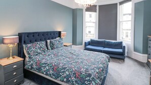 Un dormitorio con una cama de cabecera capitonada, ropa de cama floral, un sofá azul, una araña y ventanas grandes.
