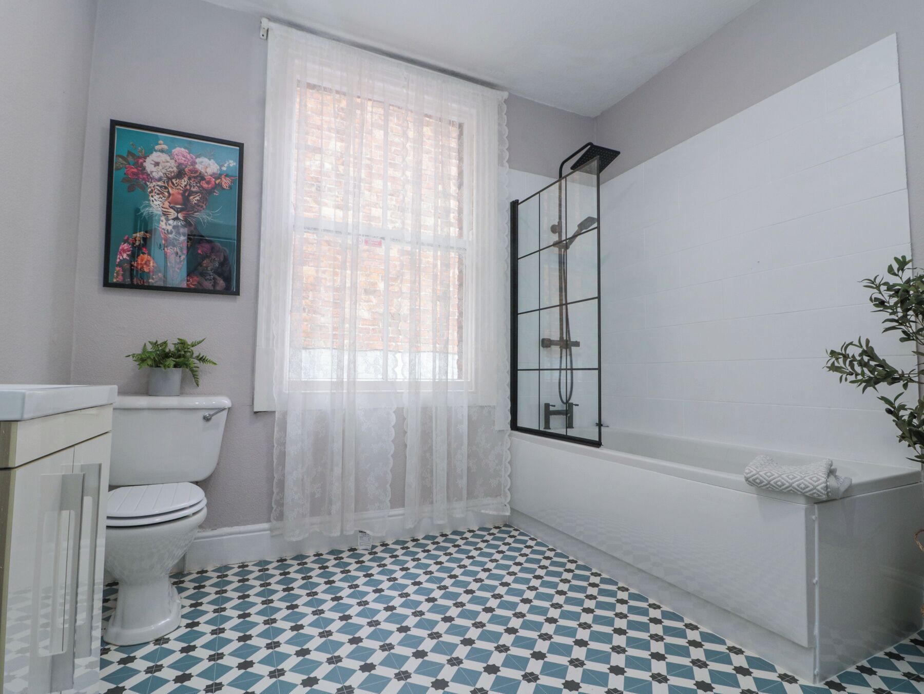 A bathroom with a toilet, a shower, a patterned floor, a window with sheer curtains, and a framed picture on the wall.