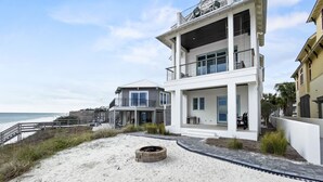 A modern beachfront house with a large balcony, a fire pit, and a view of the ocean.