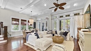 Un salon spacieux avec un grand canapé blanc, une table basse et un ventilateur de plafond. De grandes fenêtres sont agrémentées de rideaux, et on y trouve un coin repas avec une table et des chaises, ainsi qu’un escalier avec des rampes en bois.