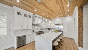 A modern kitchen with white cabinetry, a marble island, and a wooden ceiling. - Summer Wind 7 Bedroom Home by RedAwning (Santa Rosa Beach)