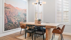 A dining area with a wooden table, black chairs, a hanging light fixture, and a large landscape painting on the wall.