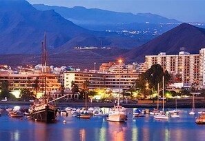 A marina with boats, a large building with multiple floors, and mountains in the background.