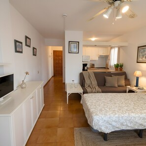 A bedroom with a bed, a TV, a desk, and a kitchenette in the background.