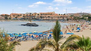 Una playa con tumbonas y sombrillas, un promontorio rocoso en el agua y edificios al fondo.
