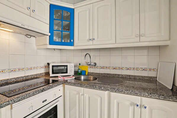 Una cocina con armarios blancos, un ventanal con marco azul, un microondas, un fregadero y una estufa.
