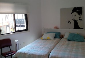 Una stanza con due letti, una sedia, un termosifone, una finestra con le sbarre e una mensola a muro con un quadro.