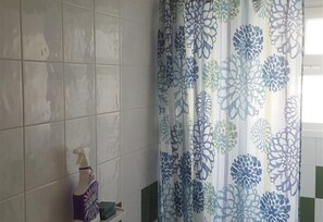 Une salle de bain aux murs carrelés, avec un rideau de douche orné de motifs floraux et une bouteille de savon liquide posée sur le sol.