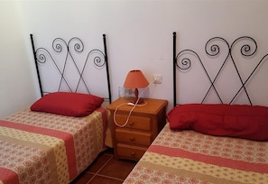 Une chambre avec deux lits simples, chacun agrémenté d’un oreiller rouge et d’une couvre-lit à motifs. Une table de chevet en bois avec une lampe se trouve entre les deux lits.