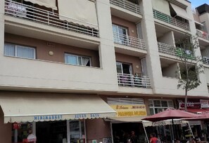 A multi-story building with balconies, a visible storefront with a sign for a bakery, and a tree in front. - Holiday Accommodation "Nice stay in Plaza Los Cristianos centre"  (Los Cristianos)