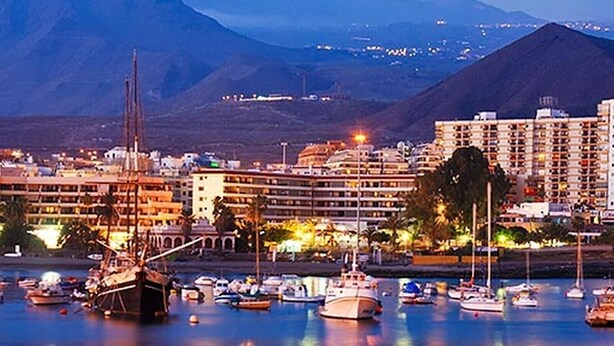 A marina with boats, a building with multiple floors, and mountains in the background.