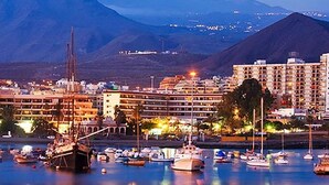 Un puerto deportivo con barcos, un edificio de varios pisos y montañas al fondo.