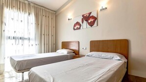 Quarto de hotel com duas camas, uma janela grande com cortinas, uma obra de arte na parede e uma mesa de cabeceira com abajur.