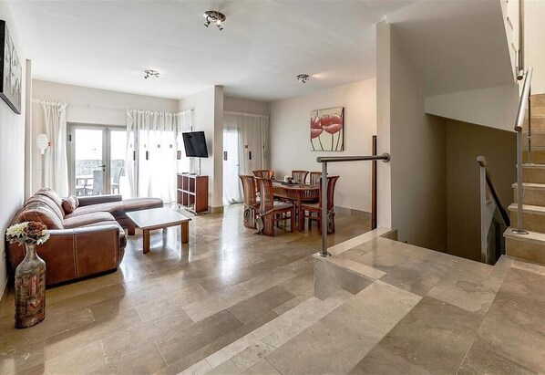 A spacious living room with a sofa, dining table, and chairs, a staircase, and a TV on a cabinet.