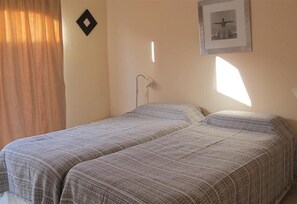 Una habitación con dos camas, una lámpara de pared, un cuadro enmarcado y una ventana con cortinas.