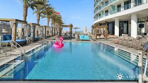 A pool area with lounge chairs, a pink flamingo float, palm trees, and a modern building with balconies. - Ocean View Balcony|Beachfront Resort|Pools+ Gym (Hollywood)