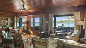 A cozy room with wooden walls, a ceiling fan, and large windows offering a view of the water. There are several chairs and a sofa arranged for seating. - Classic Walloon Lake Cottage (Boyne City)