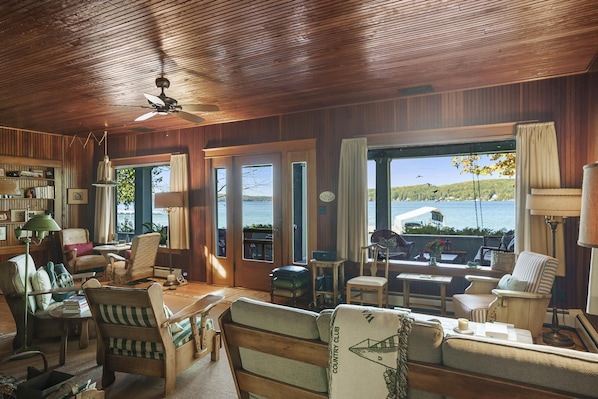 A cozy room with wooden walls, a ceiling fan, and large windows offering a view of the water. There are several chairs and a sofa arranged for seating. - Classic Walloon Lake Cottage (Boyne City)
