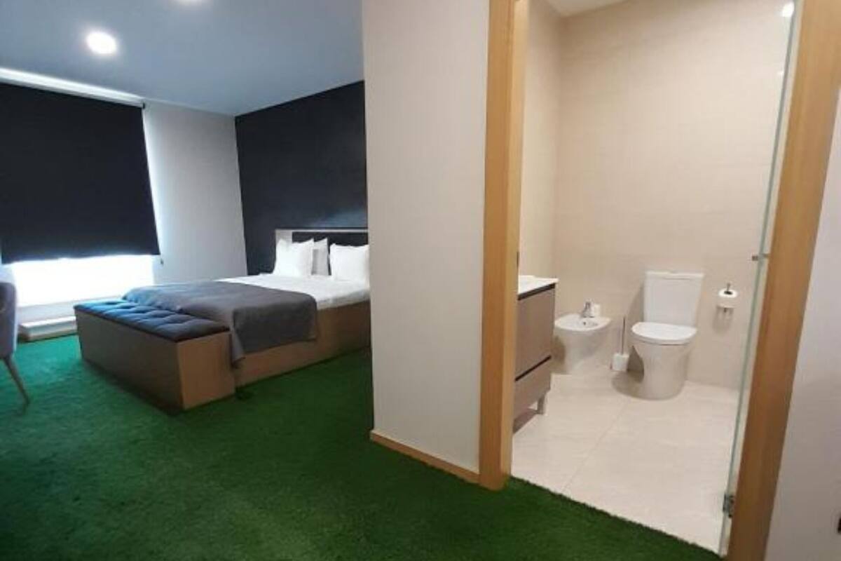 A modern hotel room with a bed, a small bedside table, a nightstand, a window with blinds, and a bathroom with a toilet, sink, and shower.
